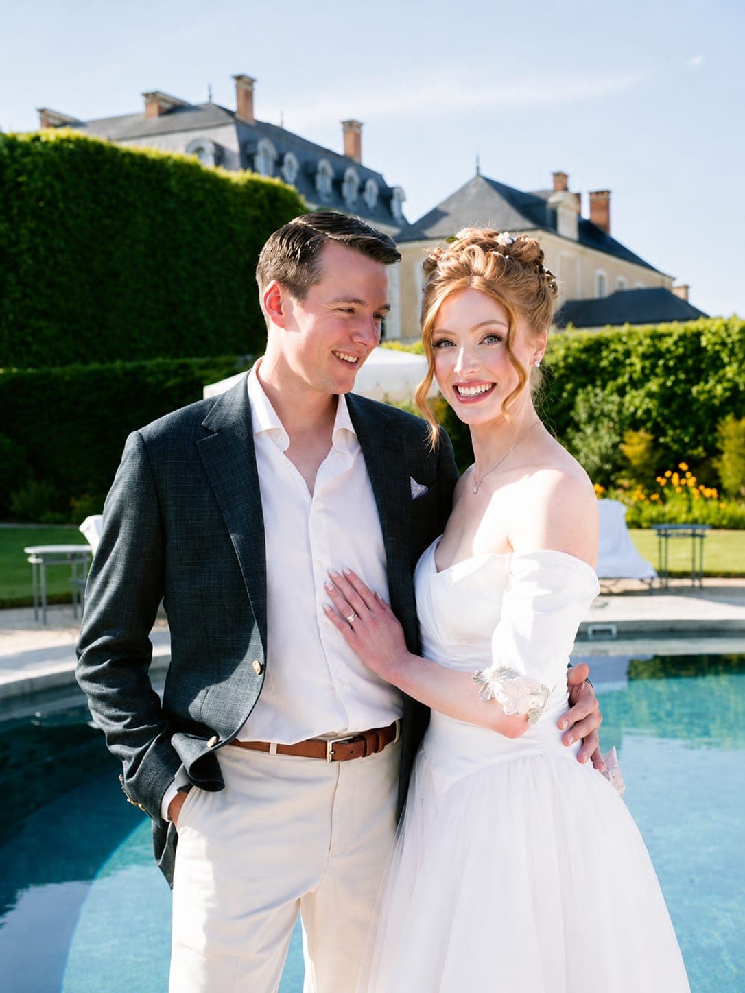 A bride and groom beside a château pool, the groom in a grey checked jacket and cream trousers