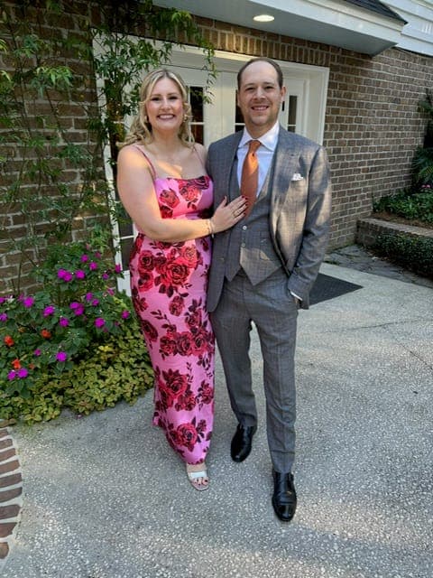 A couple outdoors before a brick wall, she in a pink floral gown, he in a grey suit