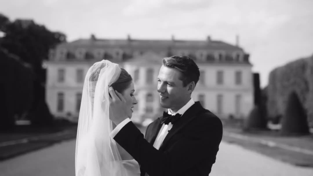 In black and white, the groom in black tie cups the bride's face, the château behind them