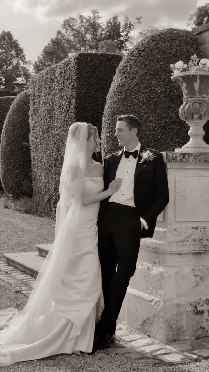 A bride in an off-the-shoulder gown and long veil rests her hand on the chest of a man in black tie with a white boutonniere, and the two smile at each other. He leans against a stone plinth topped by an urn of flowers, one hand in his pocket, with clipped topiary hedges behind. Sepia-toned black and white.