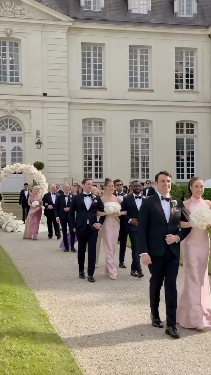 A wedding party walks toward the camera along a gravel path in front of a pale stone château — men in black tie arm in arm with women in pale pink gowns holding white bouquets, older guests following past a white floral arch